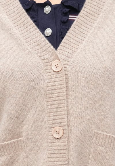 Μπεζ πλεκτή ζακέτα με δύο ανοικτόχρωμα κουμπιά πάνω από μπλε μπλούζα με βολάν στο γιακά και κουμπιά διακοσμημένα με πέρλες ορατά στη λαιμόκοψη.