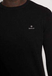 Zwarte gebreide trui met een ronde halslijn. Heeft een klein wit logo van "GANT" op de borst. De structuur lijkt geribbeld.