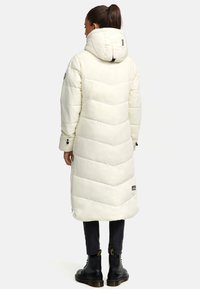 Lang, off-white pufferjakke med hætte, der har chevronmønster søm, lynlåsdetaljer og sorte accenter. Bæret med mørke bukser og støvler.