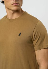 Tricou din bumbac maro cu gât rotund, mâneci scurte și un mic logo brodat negru pe piept. Textură fină și croială ajustată.