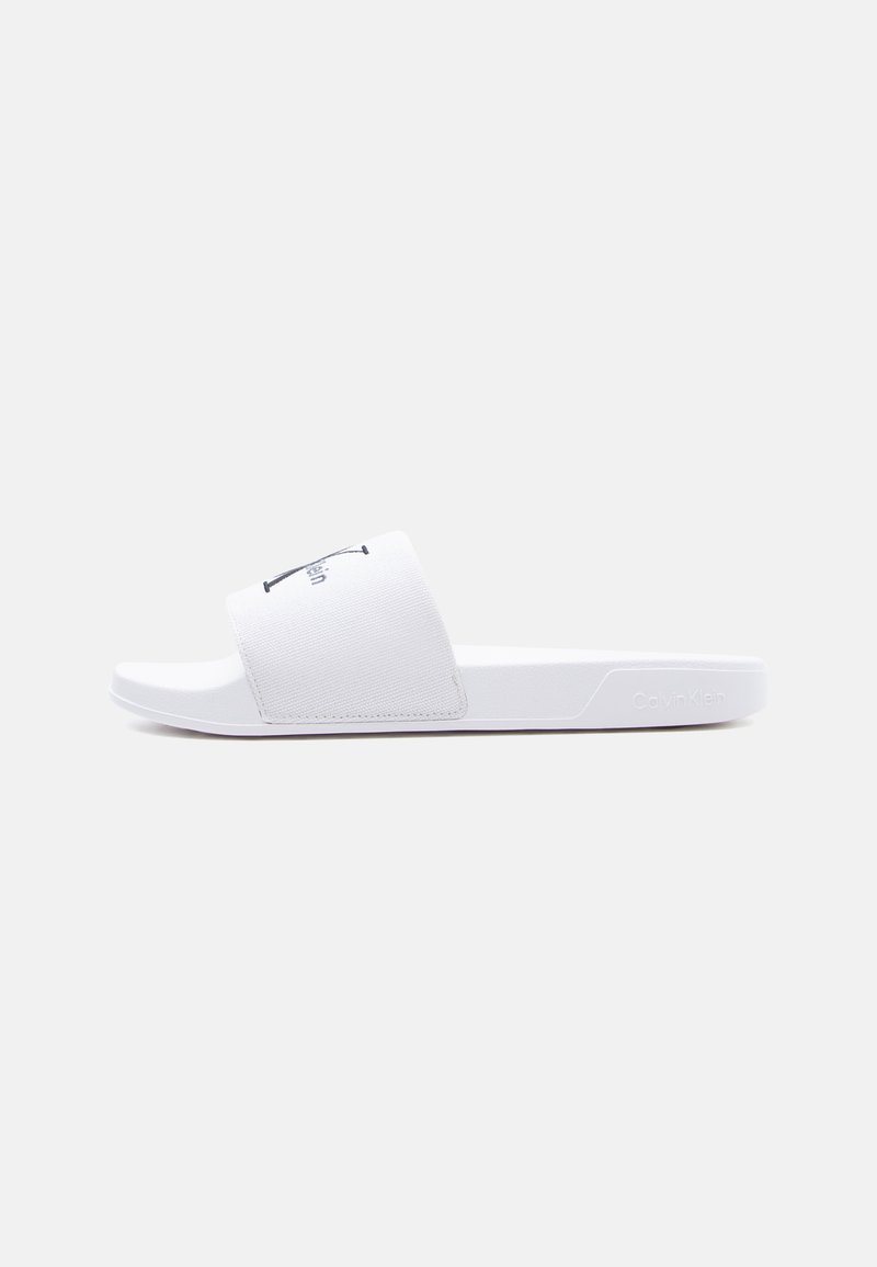 Sandale slide blanche Calvin Klein avec large bande en tissu et semelle moulée, présentée sur fond blanc uni.