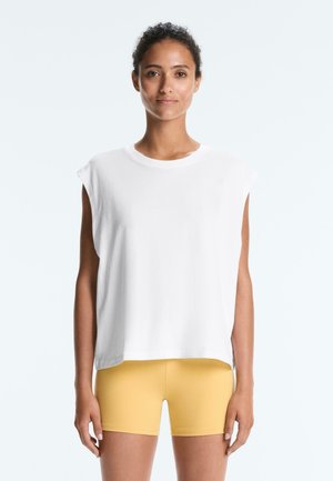 Žena s kučeravými vlasmi nosí biely top bez rukávov a žlté cyklistické šortky, stojí čelom vpred na neutrálnom svetlom pozadí.