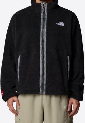 Mand iført sort fleecejakke med hvidt mønstret lynlås og beige bukser, begge med The North Face-logo.