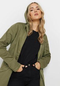 Giacca verde oliva con cappuccio e bottoni frontali, tessuto strutturato, tasche laterali, indossata sopra una camicia nera e pantaloni.