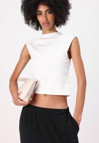 Top blanco corto con un escote alto y mangas cortas, combinado con pantalones negros. Sosteniendo un pequeño bolso beige texturizado.