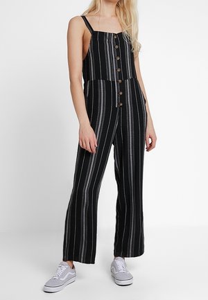 Černobílé pruhované overal s knoflíky vpředu, širokými nohavicemi a nastavitelnými ramínky, doplněný bílými teniskami.