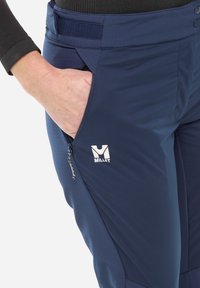 Pantalons de plein air bleu marine avec une poche zippée et une taille élastique. Présence d'un logo Millet blanc près de la poche. Texture lisse.