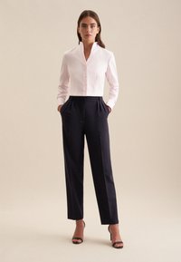 Camicia collare rosa chiaro con bottoni, abbinata a pantaloni a pieghe blu navy scuri e sandali neri con tacco alto, su uno sfondo neutro.