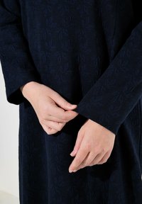 Indumento di un blu scuro con una texture che presenta un motivo a foglie, mentre una persona sta chiudendo l'apertura della manica con entrambe le mani.