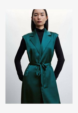 Satin-türkis ärmelloser Trenchcoat mit Schalkragen, tailliertem Gürtel und glatter Textur, getragen über einem enganliegenden schwarzen Langarmoberteil.