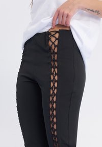Pantalon noir avec un design à lacets sur les côtés, une texture lisse et une coupe ajustée qui dévoile la peau sous la dentelle.