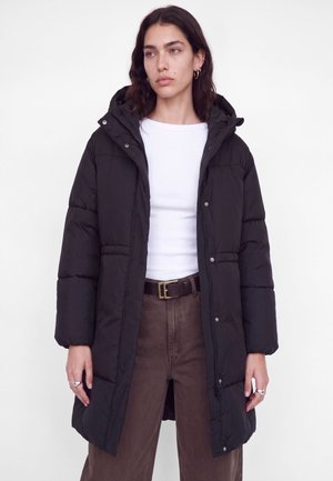 Une femme présente un long manteau matelassé noir à capuche, porté sur un haut blanc et un pantalon marron, devant un fond de studio uni.