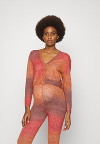 Conjunto de cardigan em malha e leggings em tons de laranja degradê. Apresenta um decote em V, mangas longas e fecho central com botões. Tecido macio e texturizado.