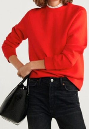 Kobieta w jasno czerwonym swetrze z długim rękawem, ciemnych, wysokich dżinsach i trzymająca czarną teksturowaną torebkę na tle jednolitego tła.