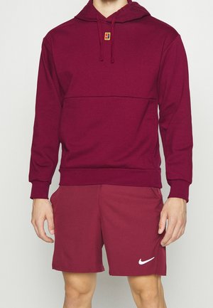 Mężczyzna w bordowej bluzie z kapturem z małym kolorowym logo na piersi oraz pasujących bordowych szortach Nike z białym znakiem Swoosh na lewej nodze.