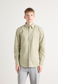 Camisa de botones verde oliva claro con mangas largas, que presenta un cuello clásico, bordado sutil y un corte relajado. Combinada con pantalones grises.