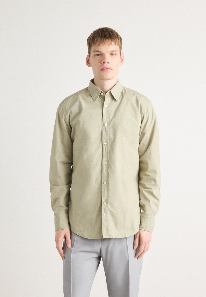 Camisa de botones verde oliva claro con mangas largas, que presenta un cuello clásico, bordado sutil y un corte relajado. Combinada con pantalones grises.