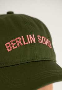 Căciulă olive cu text brodat roz "BERLIN SOHO." Fabricată din material textil, cu margine curbă și design structurat.