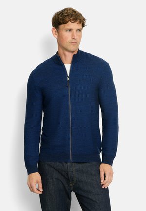 Dunkelblaues, mit Reißverschluss versehenes Strick-Cardigan mit strukturiertem Muster, hohem Kragen und gerippten Bündchen und Saum. Kombiniert mit dunklen Denim-Jeans.