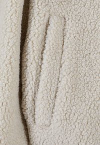 Maglione in pile bianco sporco con superficie testurizzata e soffice e dettagli di tasche laterali verticali. Materiale morbido con un aspetto accogliente.
