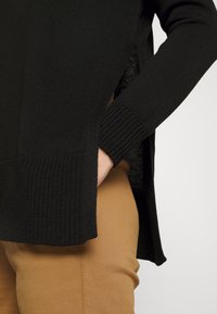 Personne portant un pull noir en maille avec des fentes sur les côtés et un pantalon couleur camel, la main glissée dans la poche du pull.