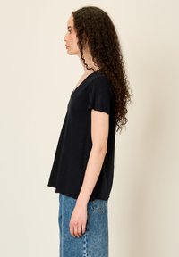 Young woman with long curly hair wearing a black short-sleeve V-neck shirt and blue jeans, standing in profile against a plain background.