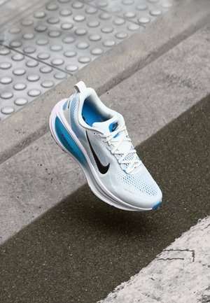 Baskets de course Nike blanches et bleues avec un swoosh noir flottant au-dessus d'un trottoir texturé et d'un bord de rue en asphalte.