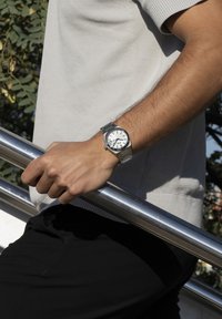 Reloj de pulsera de acero inoxidable plateado con esfera blanca, marcadores negros y pulsera de metal. El portador sostiene una barandilla de acero inoxidable.