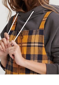 Żółto-granatowa sukienka na szelkach w kratę z przednim zamkiem błyskawicznym i regulowanymi paskami, noszona na szarej bluzie z kapturem. Tkanina o fakturze.