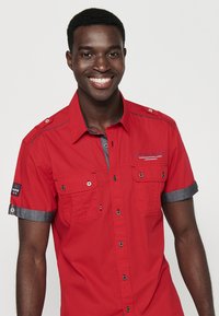 Camisa roja de manga corta con dos bolsillos frontales, detalles grises en el cuello y las mangas, estilo de botones, con un logotipo en la manga derecha.