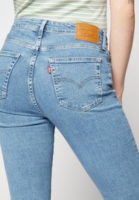 Personne portant un jean Levi's bleu clair et une chemise rayée, montrant les poches arrière et la patch en cuir de la marque sur la taille.