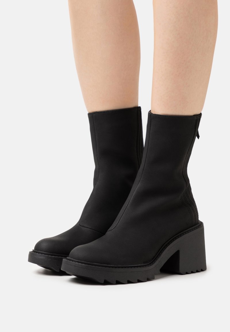 Paire de bottes noires mi-mollet avec talons hauts épais et semelles rainurées portées sur des jambes nues sur fond blanc.