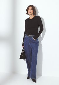 Black long-sleeve top paired with high-waisted blue jeans, a black belt, black pointed shoes, and a small black handbag against a light background.