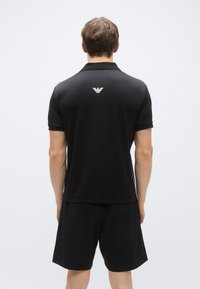 Svart poloshirt med ribbekrave og korte ermer, med en hvit logo på baksiden. Kommer sammen med matchende svarte shorts.