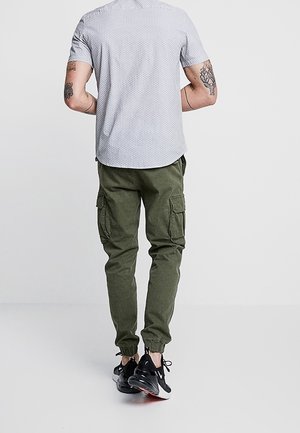 Hombre de espaldas vistiendo una camisa de manga corta estampada, pantalones cargo verde oliva y zapatillas deportivas negras.