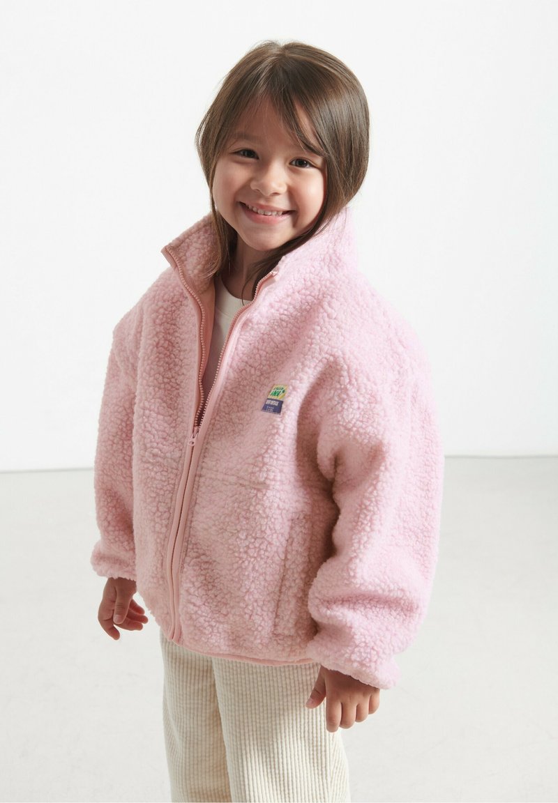 Chaqueta de forro polar rosa con cuello alto y cremallera frontal. Presenta un material texturizado y esponjoso, además de un pequeño logotipo en parche. Combinada con pantalones a rayas.