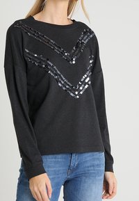 Nainen, jolla on päällään musta pitkähihainen pusero, jossa on kaksi viistosti aseteltua mustaa paljettiraidetta, yhdistettynä sinisiin farkkuihin.