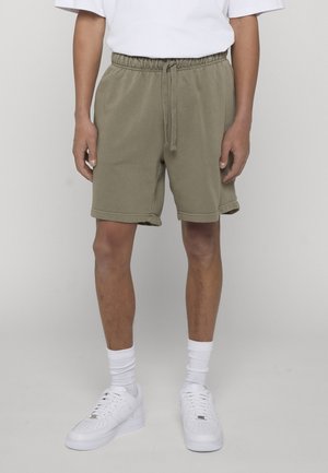 Osoba ubrana w oliwkowozielone szorty z sznurkiem, białe skarpetki za kostkę i białe trampki na szarym, jednolitym tle.