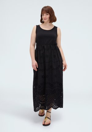 Frau mit kurzen braunen Haaren, die ein ärmelloses schwarzes Maxikleid mit ausgeschnittenen Blumenmustern und schwarze Sandalen mit goldenen Akzenten trägt.