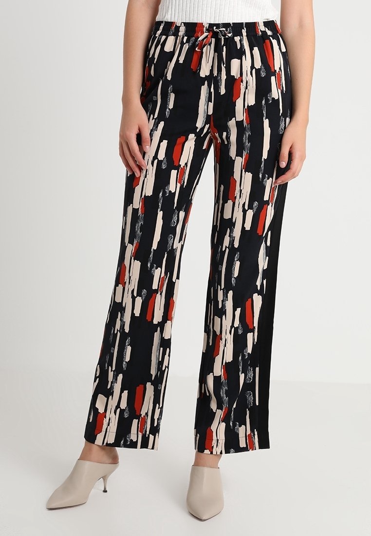 Femme portant un pantalon large noir avec des motifs de coups de pinceau abstraits rouges, beiges et blancs, associé à des mules à talons beiges.