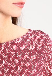 Punainen kuviollinen paita, jossa on valkoinen geometrinen kuvio, pyöreä kaula-aukko ja sileä kangas.