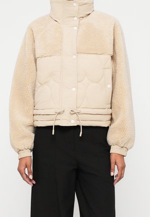 Veste courte beige en polaire et matelassée avec des boutons-pression, col montant, ourlet à cordon de serrage et poignets élastiques, portée avec un pantalon noir.