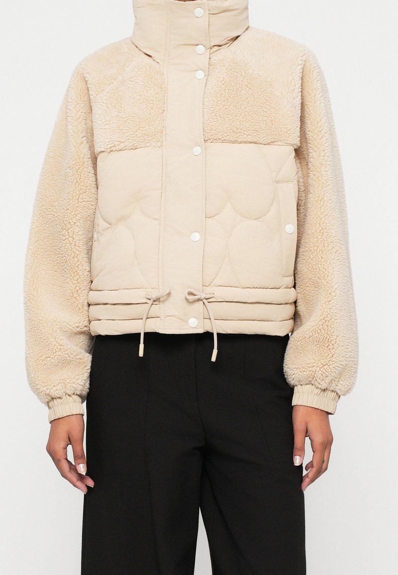 Veste courte beige en polaire et matelassée avec des boutons-pression, col montant, ourlet à cordon de serrage et poignets élastiques, portée avec un pantalon noir.