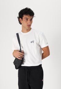 Witte katoenen t-shirt met een donker logo op de linkerborst. Gecombineerd met zwarte joggingbroeken en een kleine zwarte schoudertas.