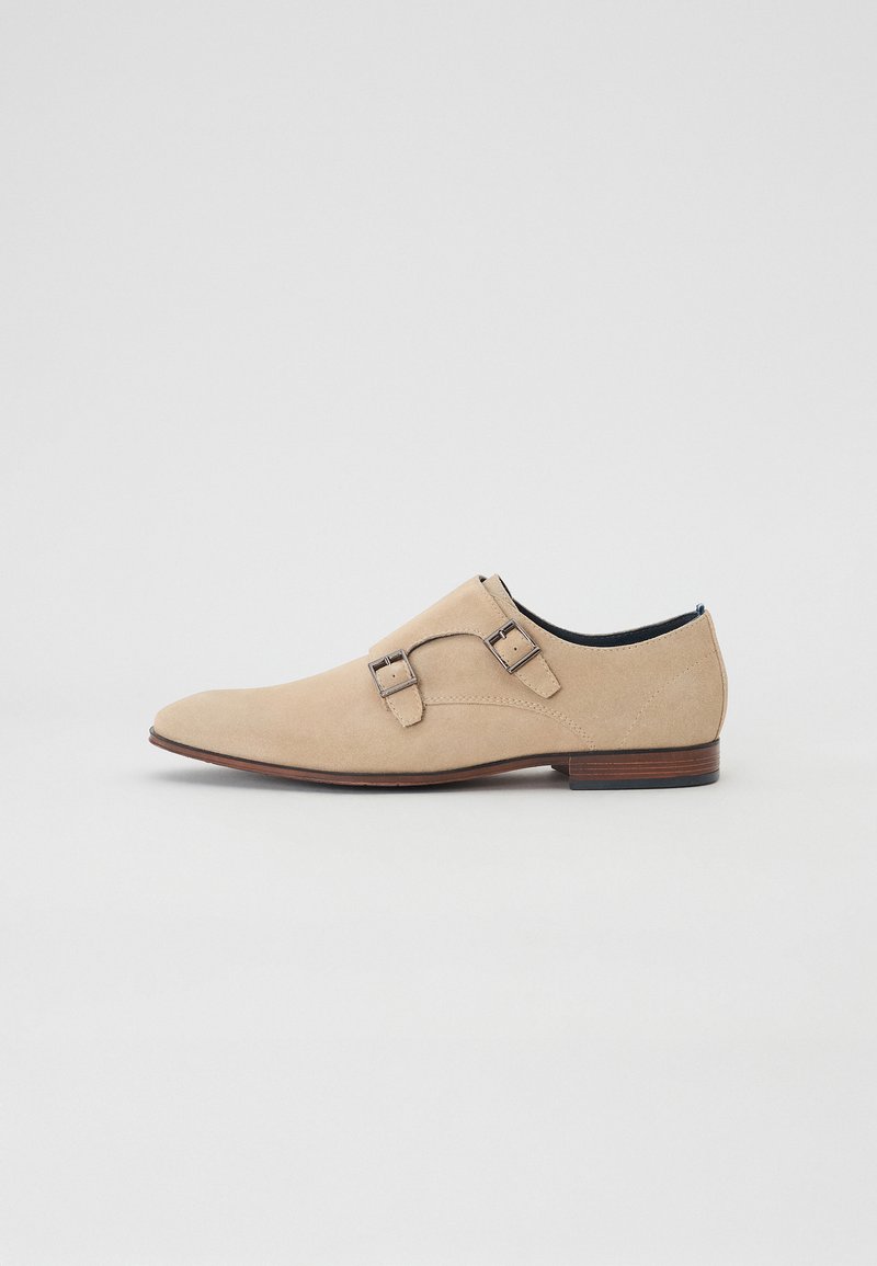 Beige Wildlederschuh mit spitzer Zehenform, doppelten Schnallenriemen und einem niedrigen Holzabsatz. Glatte Textur und minimalistisches Design.