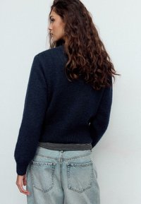 Maglione blu navy con maglia testurizzata, scollo rotondo e maniche a palloncino, abbinato a jeans in denim chiaro con una banda elastica a righe in vita.