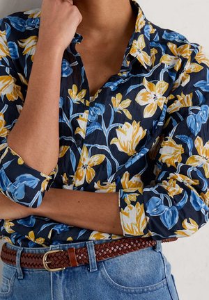 Osoba ubrana w granatową koszulę z żółto-niebieskim kwiatowym wzorem, z podwiniętymi rękawami, ręką przy brodzie oraz brązowy skórzany pleciony pasek i niebieskie dżinsy.