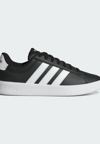 Baskets basses noires Adidas avec trois bandes blanches, semelle blanche et lacets noirs, montrées de profil sur un fond clair.