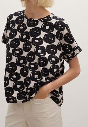 Frau trägt ein schwarz-weiß gemustertes kurzärmliges Hemd, die Hand in der Tasche einer hellen Hose, vor einem einfarbigen Hintergrund.