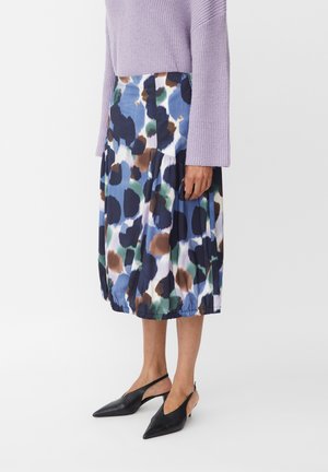 Model draagt een midi-lange rok met abstracte vlekken in blauw, bruin, zwart en groen, gecombineerd met een lavendel trui en zwarte slingback hakken.
